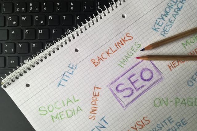Image of a notebook on top of a keyboard, with buzzwords like "SEO" and "social media" written on it with different colored pencils, with two colored pencils resting on top of the notebook.